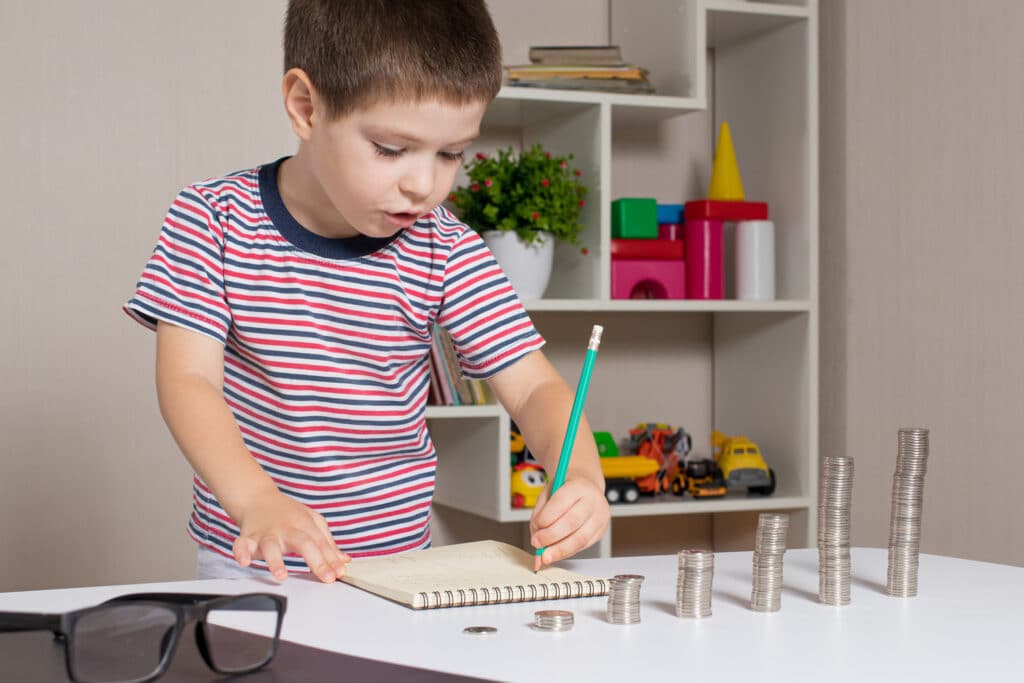 little boy writing in notebook with stacks of coins