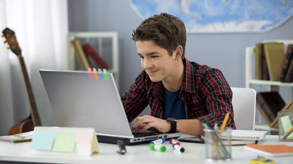 teen boy smiling as he does homework on a laptop